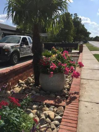 A stone planter with bright pink flowers sits in a landscaped brick garden bed next to a sidewalk and a white truck.