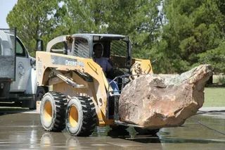 A yellow skid-steer loader carries a large, jagged brown rock on a paved surface near a white truck and green trees.