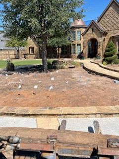 A front yard with a large tree and marking flags near a brick house, viewed from the driver's seat of a tractor.