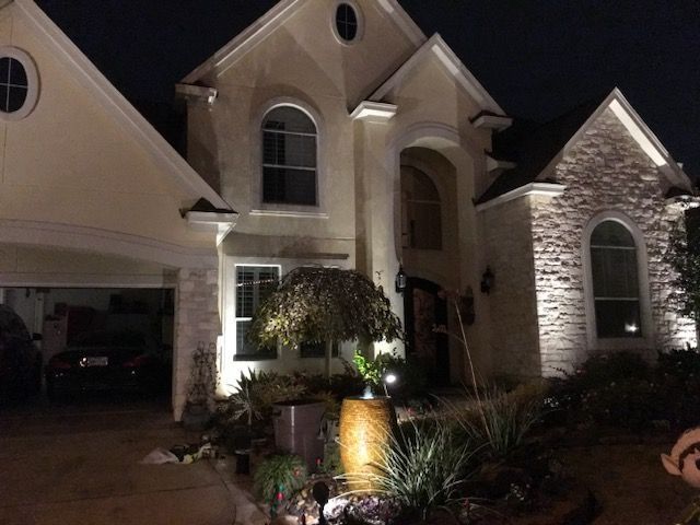 A two-story suburban home at night with exterior landscape lighting illuminating the stone facade, windows, and tree.