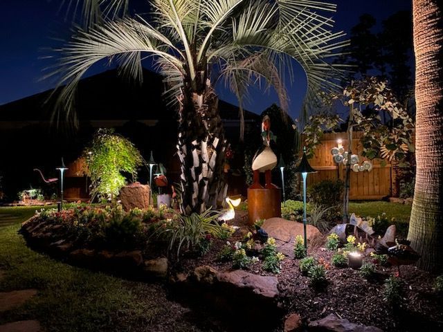 A garden at night, illuminated by small landscape lights, featuring a palm tree, stone borders, and decorative lawn ornaments.