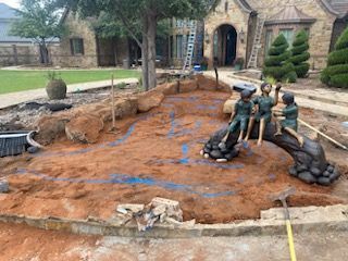 A sculpture of three figures sitting on a log centerpiece within a partially excavated, dirt-filled backyard pond area.