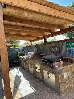 An outdoor stone kitchen with a grill and sink, sheltered by a wooden pergola against a corrugated metal fence.