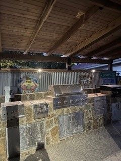 Outdoor kitchen with a built-in stainless steel grill, stone base, and corrugated metal backsplash under a wood ceiling.