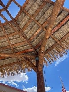 Low-angle view of a wooden thatch-roofed umbrella against a bright blue, partly cloudy sky.