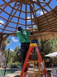 A person in a green shirt and dark pants stands on an orange ladder, repairing the thatched roof of an outdoor gazebo.