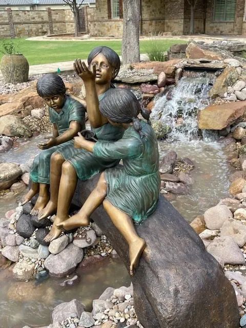 Bronze statues of three children sitting on a log by a small stone water feature in a garden.