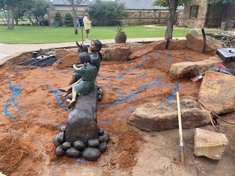 A bronze sculpture of two figures sitting on a stone log amidst a landscaped area with dirt, rocks, and a tool.