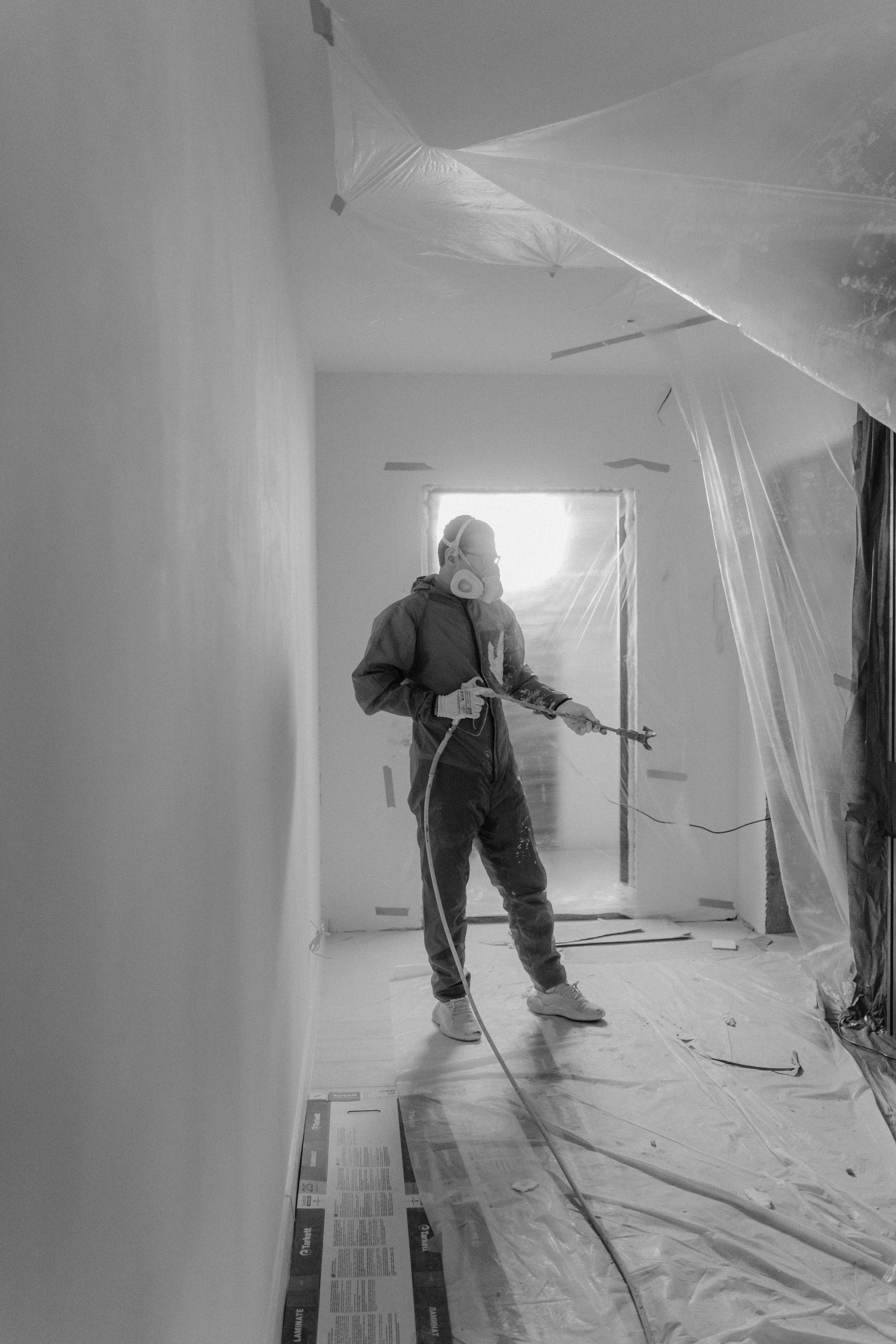 Person in protective gear spraying paint inside a room. Plastic sheeting covers surfaces; a hallway is visible.