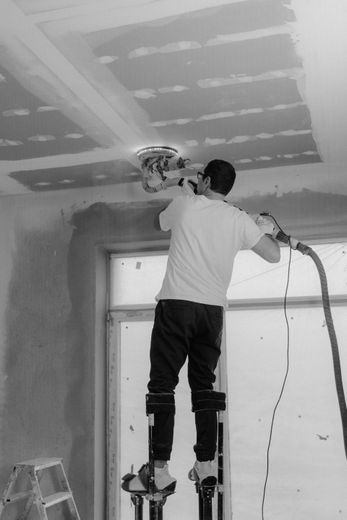 Man sanding a ceiling on stilts; dust hose connected to sander. Interior setting.