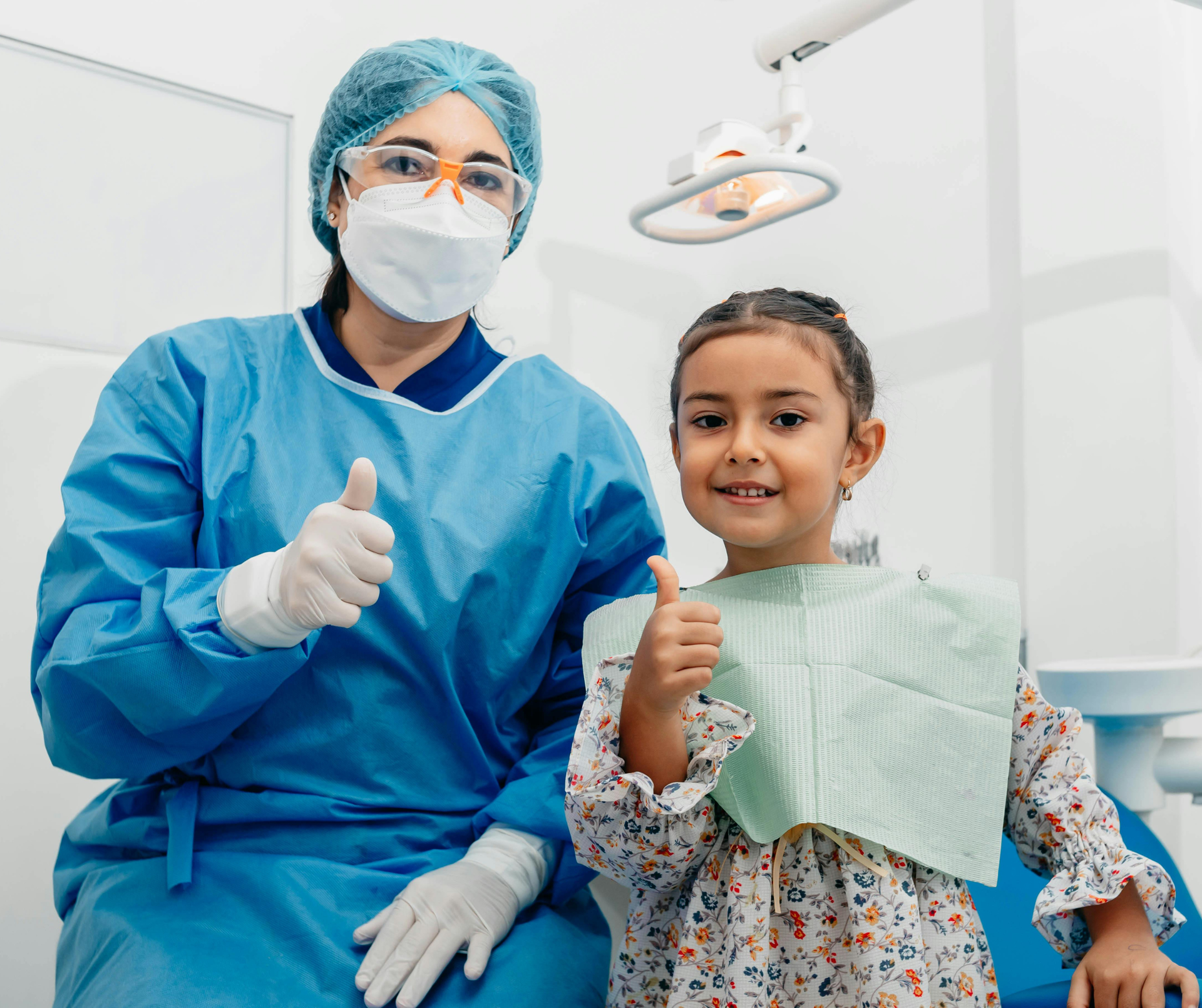 Dentist in blue scrubs and young patient in dental bib giving thumbs up in a dental office.