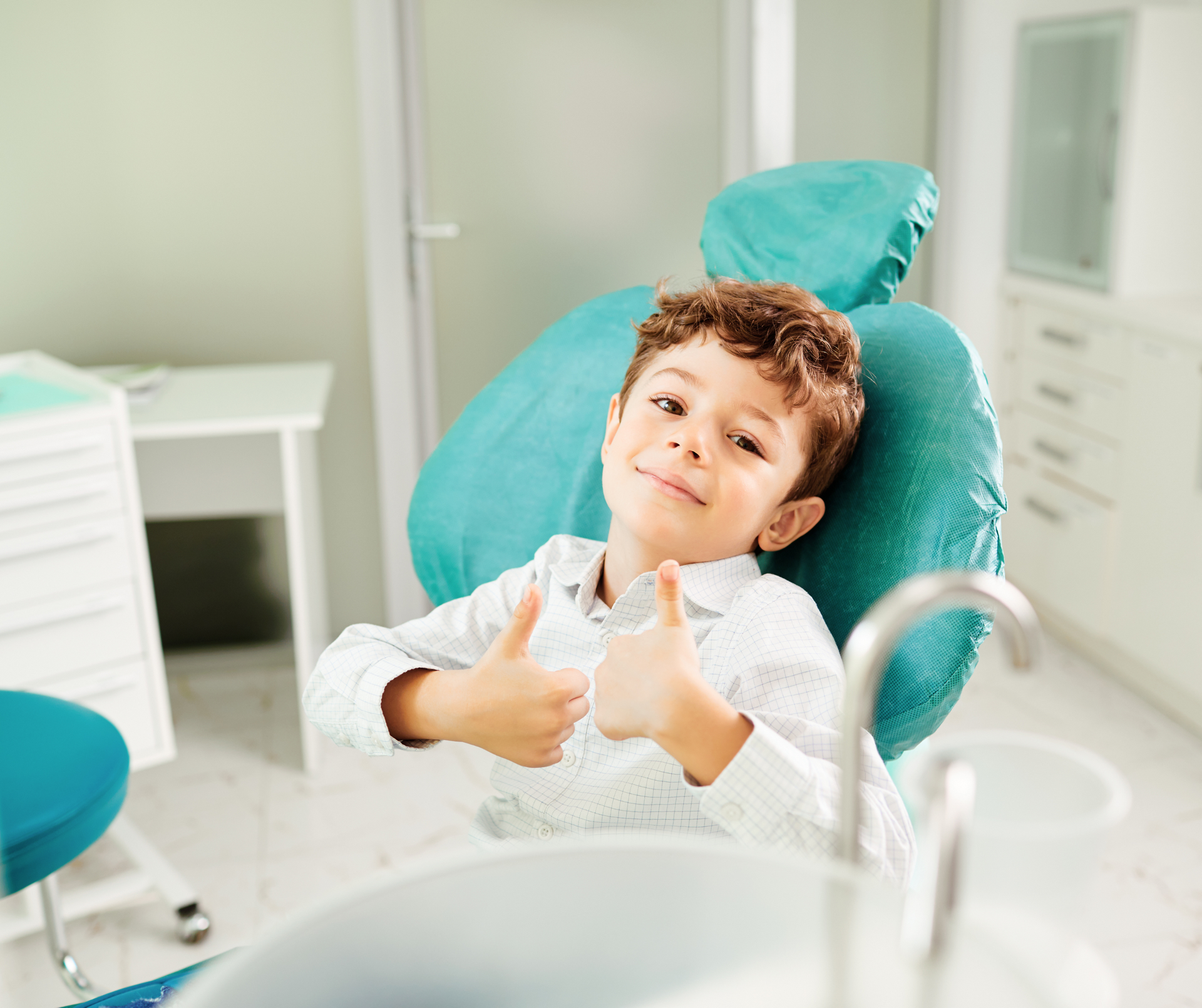 Boy in a dental chair gives thumbs up, smiling. Teal chair and medical setting.