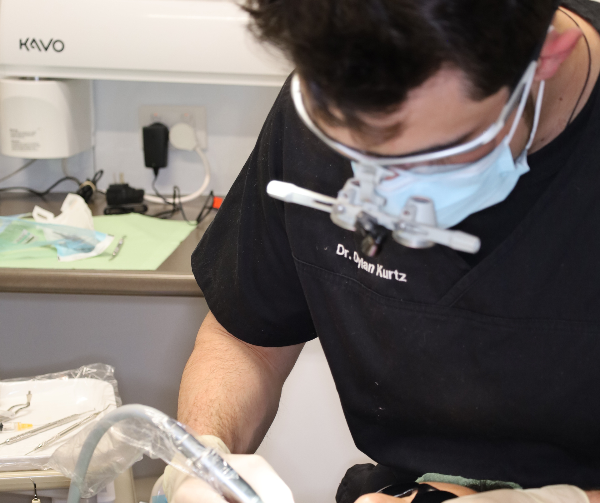 Dentist wearing loupes and mask, leaning over a patient in a dental setting.