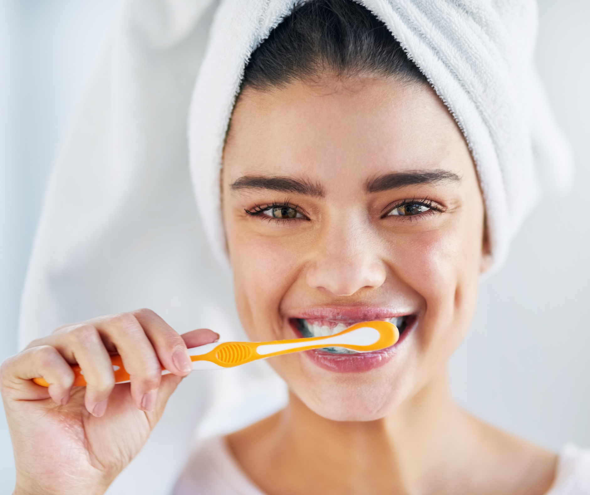 Woman brushing teeth with an orange toothbrush, wearing a white towel on her head, smiling.