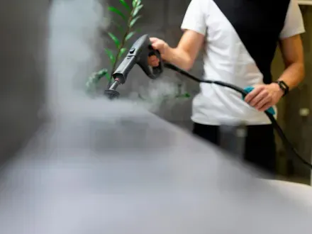Una persona con una camisa blanca y negra utiliza una limpiadora a vapor portátil, liberando una nube de vapor.