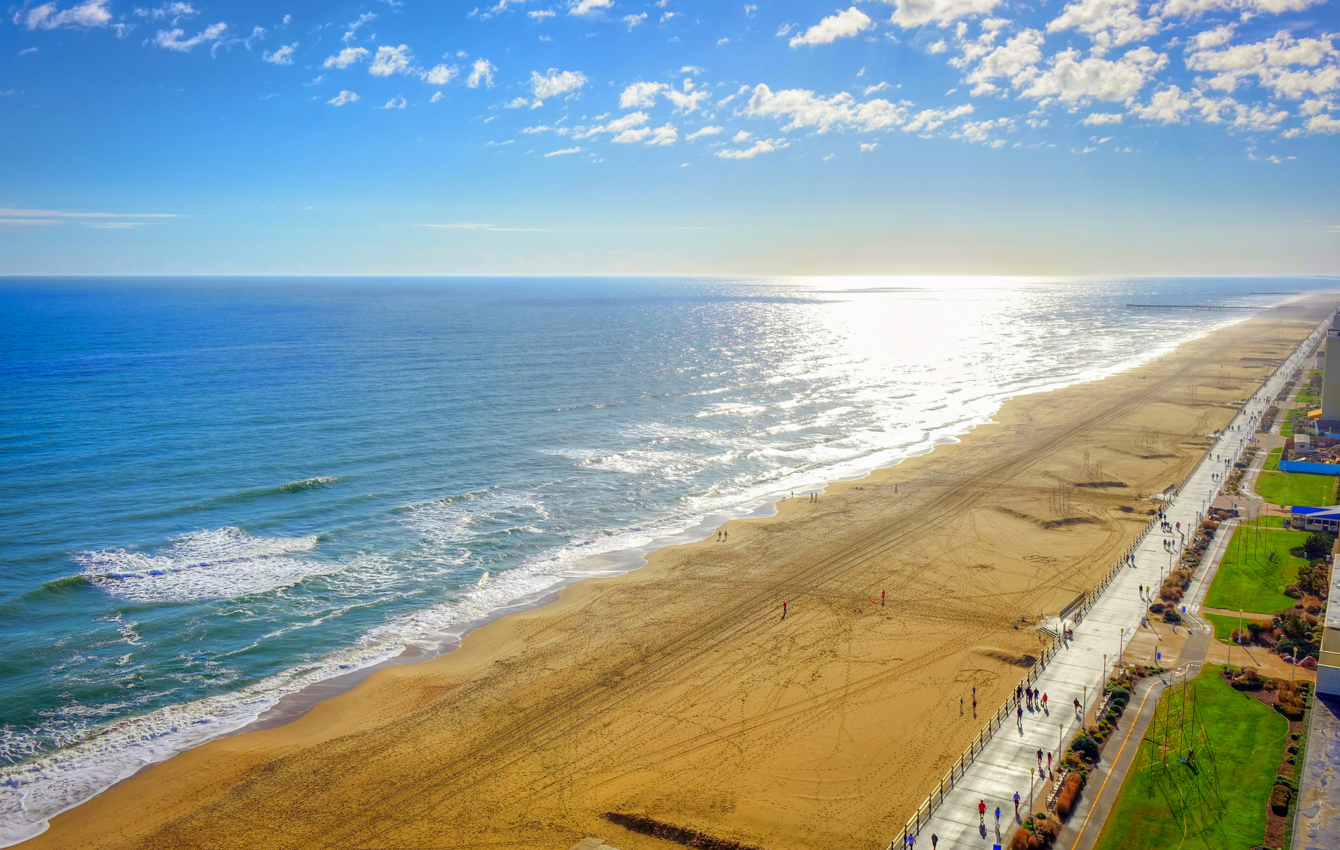 Sandy beach with ocean waves under a bright blue sky, with a road and grassy area along the shore.