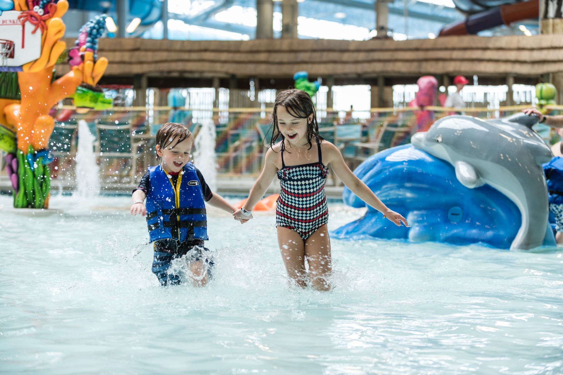 Two children run holding hands in a water park, water splashing.