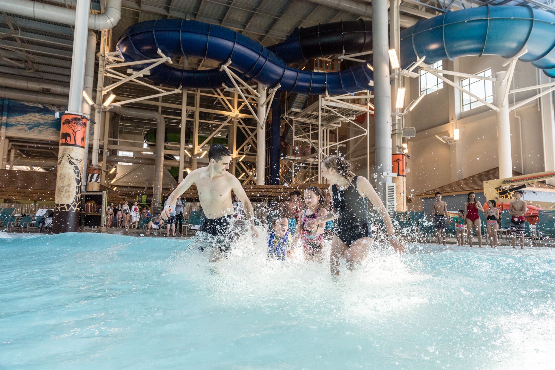 People run into a wave pool at an indoor water park; blue water, large slides visible overhead.