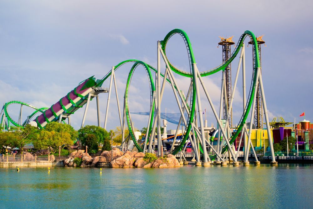 Green roller coaster with loops and twists, set over water.
