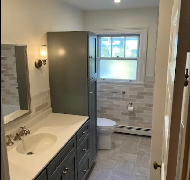 Gray bathroom with a vanity, tall cabinet, toilet, and window.