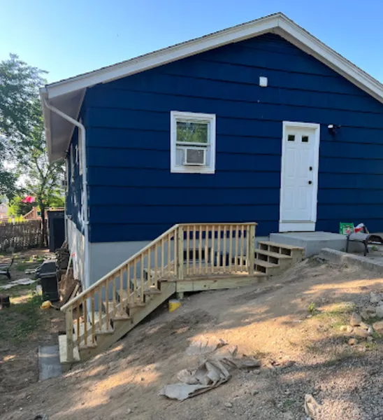 Blue house with wooden stairs leading to a white door. A window with an AC unit is also visible.