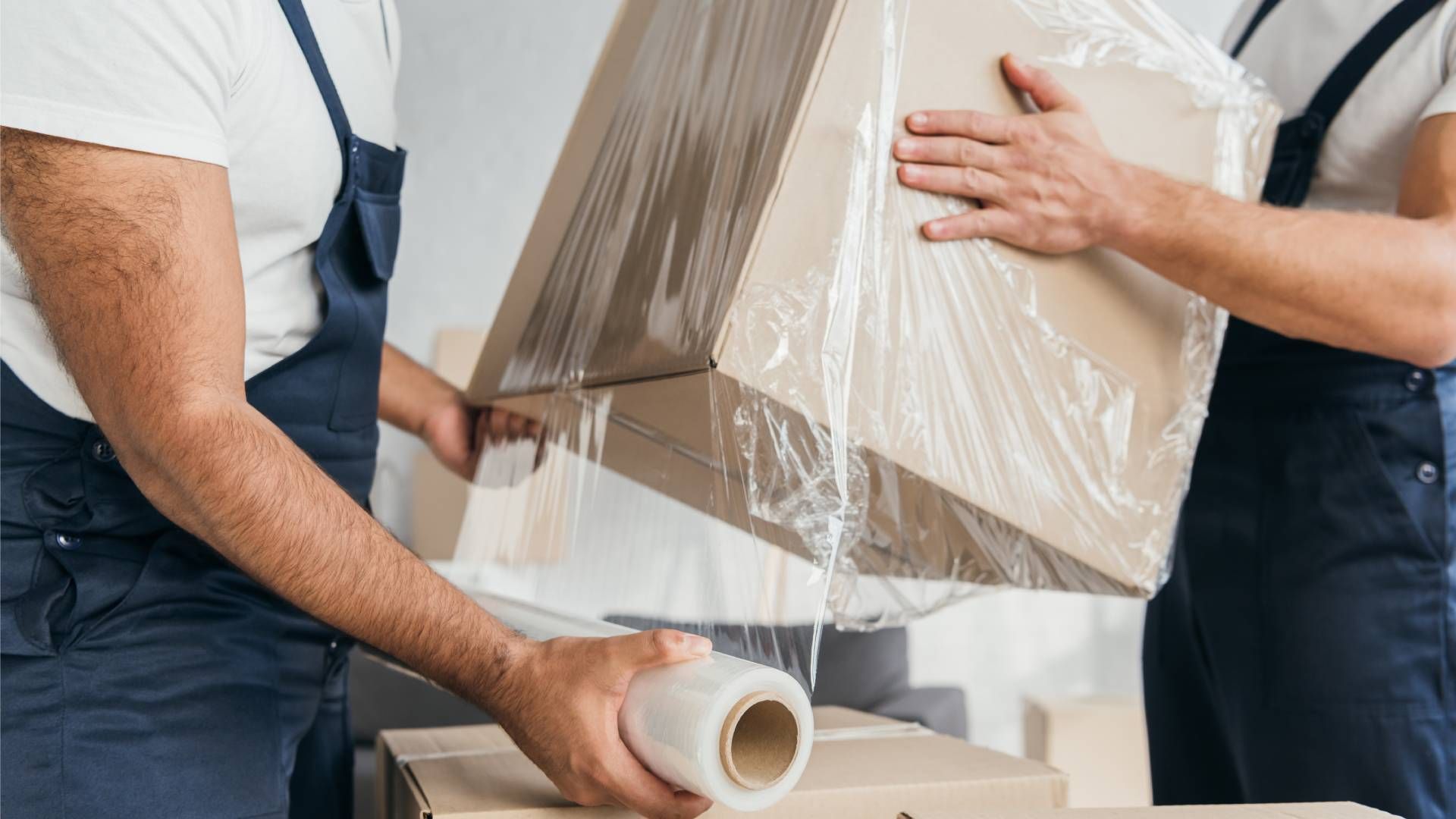 Two movers wrapping a cardboard box with clear plastic wrap.