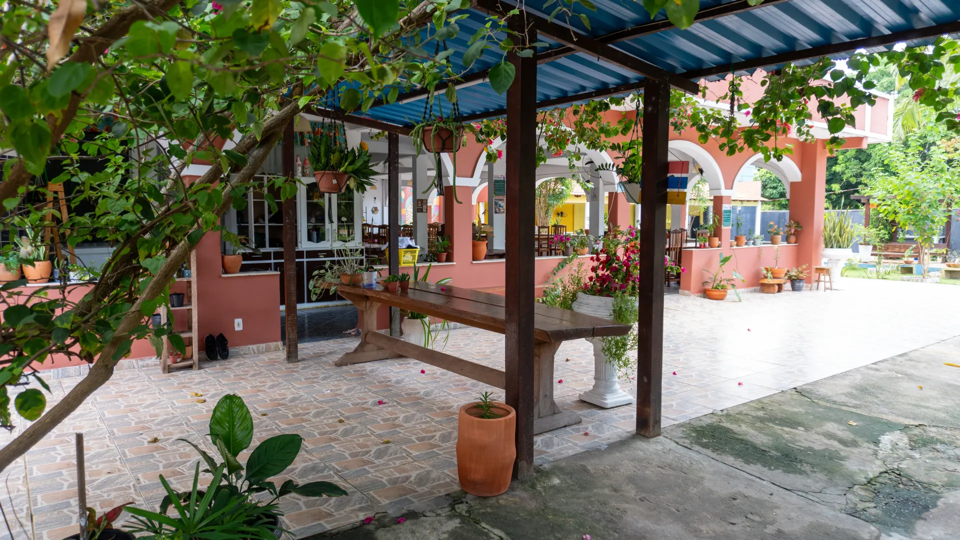 Um pátio externo coberto com uma mesa de madeira, plantas e um prédio vermelho ao fundo -Solar Café e Pousada
