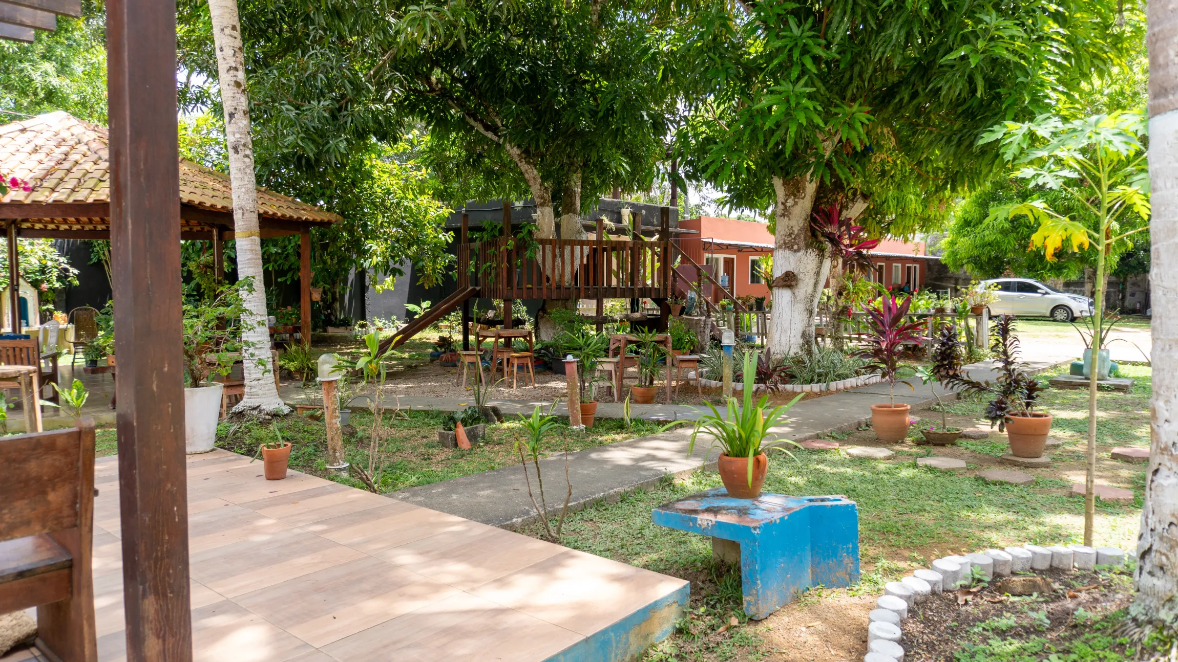 Um quintal exuberante com um gazebo de madeira, uma casa na árvore e canteiros. Verde vibrante e ensolarado com detalhes em azul - Solar Café e Pousada