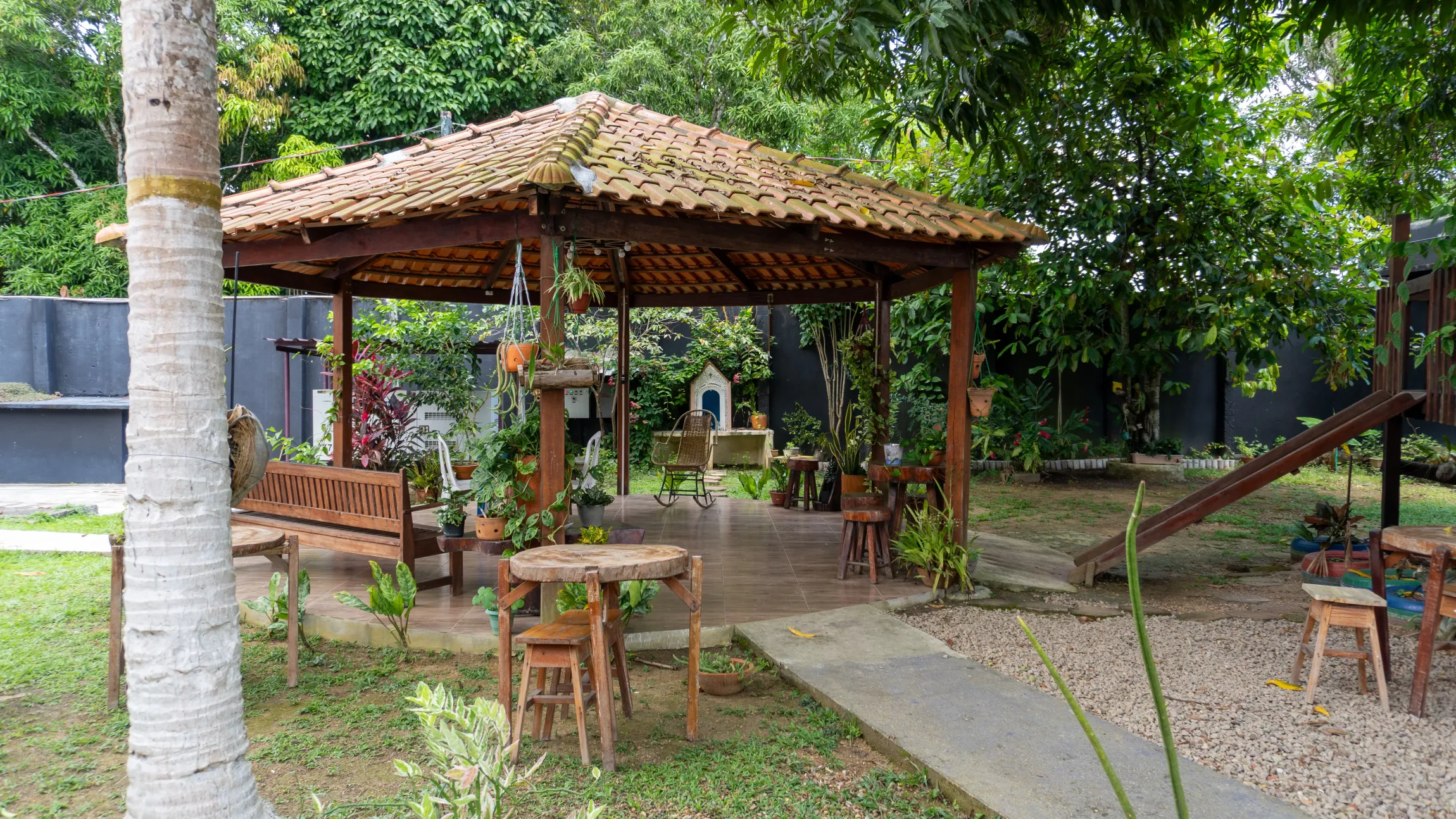 Um gazebo rústico com móveis de madeira fica em um jardim com árvores - Solar Café e Pousada