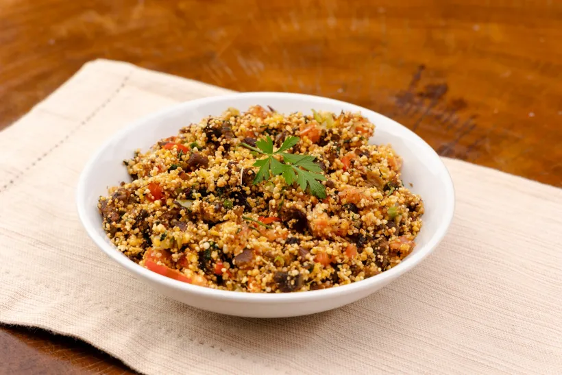 Tigela branca de quinoa temperada com tomates vermelhos e ervas verdes sobre um jogo americano bege e mesa de madeira - Solar Café e Pousada