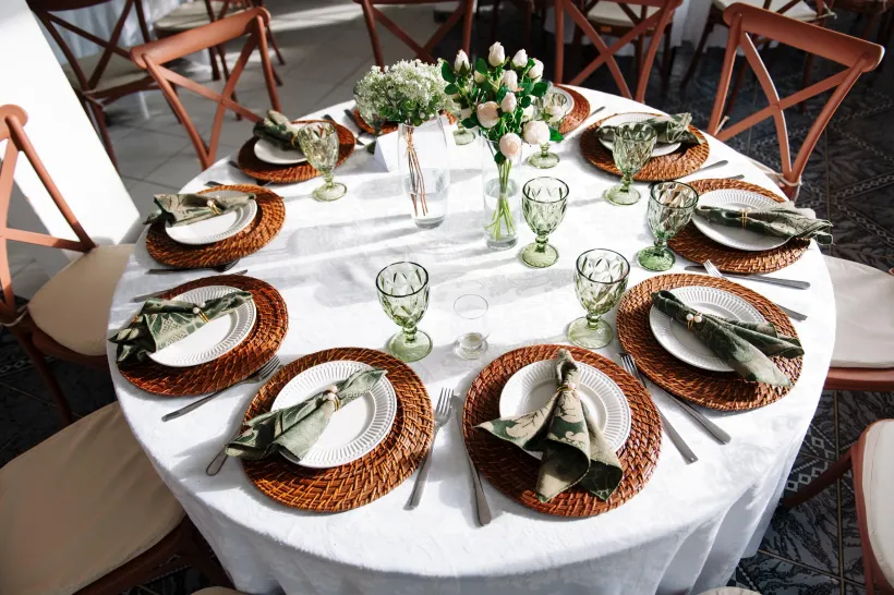 Mesa redonda posta para refeição com jogos americanos de tecido, copos verdes e centro de mesa floral - Solar Café e Pousada