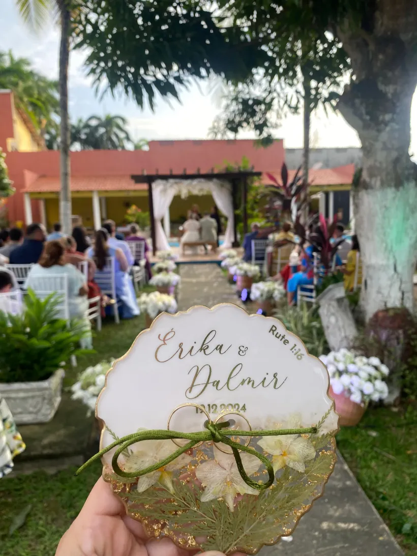 Convite para cerimônia de casamento em primeiro plano; casal no altar sob o gazebo, convidados sentados. Folhagem verde, detalhes dourados, cenário externo - Solar Café e Pousada