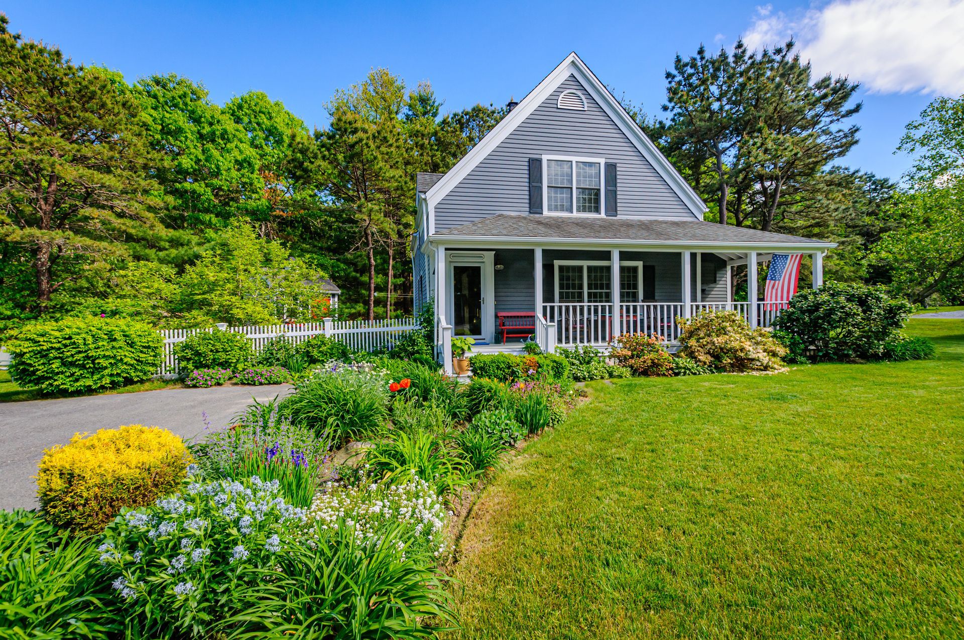 Gray house with white porch and American flag, surrounded by flowers and green lawn.