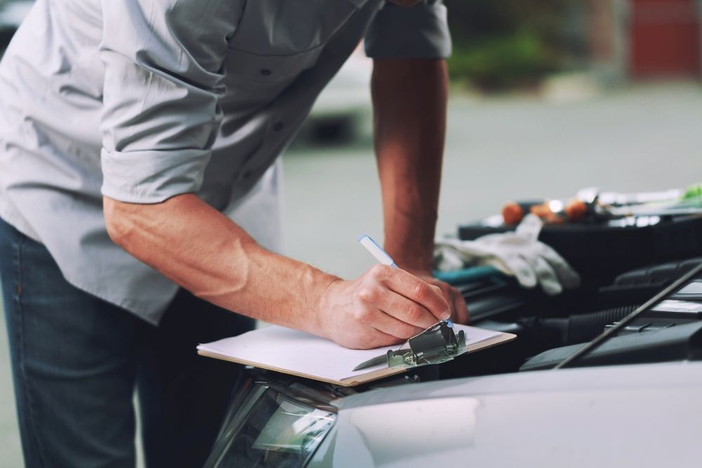 A Man is Writing on a Clipboard While Looking Under the Hood of a Car — TeeJay Mechanical Repairs In Queanbeyan, NSW