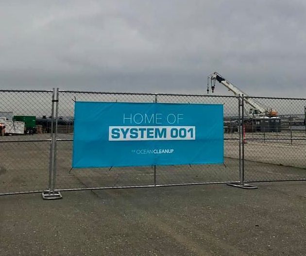 A blue sign on a chain link fence says home of system 001