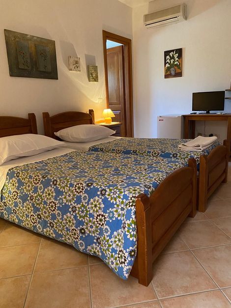 Camera da letto con due letti in legno, copriletto blu a fiori, pavimento piastrellato, quadri alle pareti, lampada e scrivania con TV.