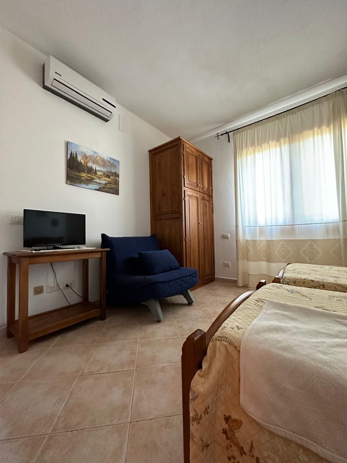 Camera da letto luminosa con letto, poltrona blu, TV su supporto in legno, armadio e finestra soleggiata.