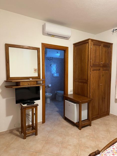 Piccola stanza con mobili in legno, specchio a parete, mobile porta TV, condizionatore e porta aperta su un bagno illuminato di blu.