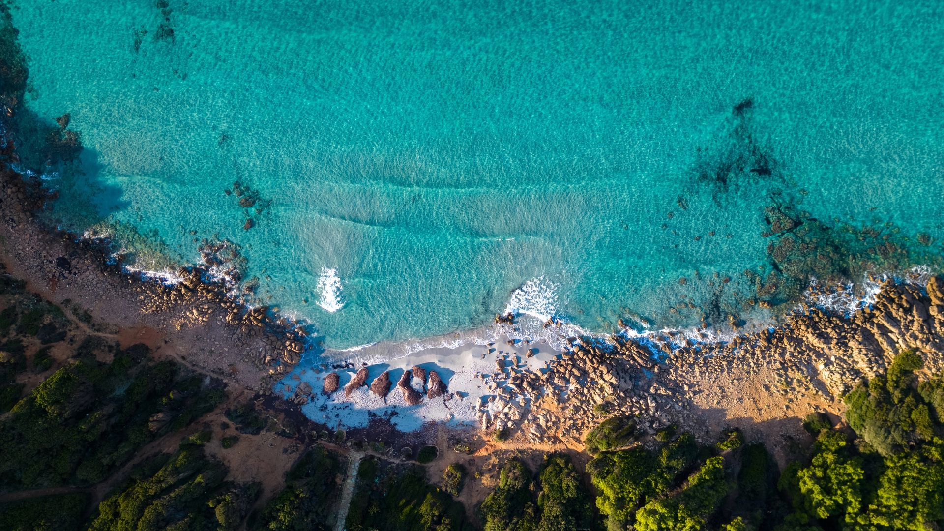 Veduta aerea di una spiaggia turchese con onde, costa rocciosa e fitta vegetazione.