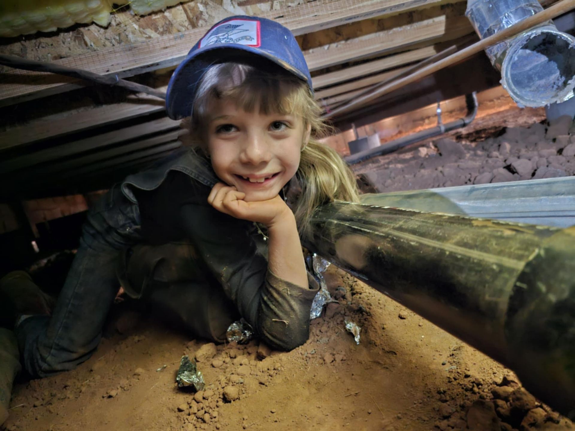 A little girl wearing a hard hat is sitting under a pipe.