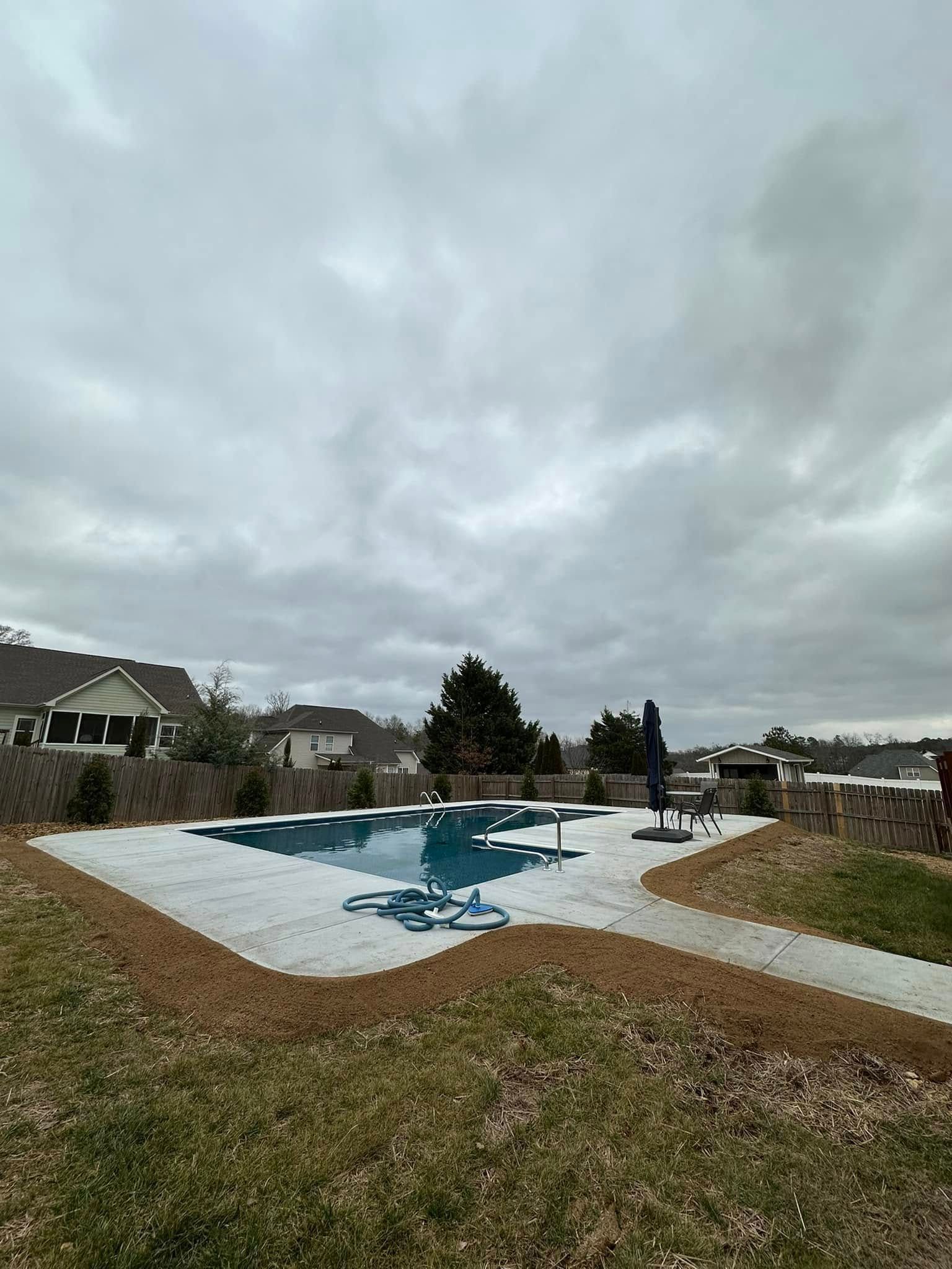 An outdoor swimming pool surrounded by a stone patio and mulch, set against a cloudy sky and neighborhood homes.