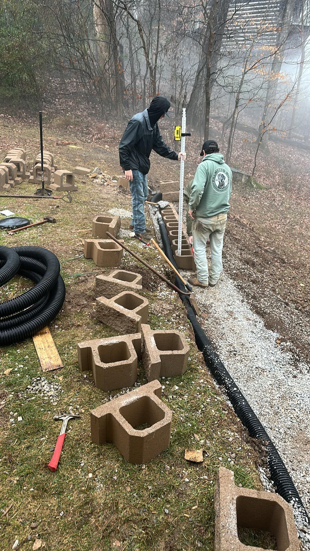 Two people work on a stone retaining wall in a yard, installing a vertical support pipe amid concrete blocks and gravel.