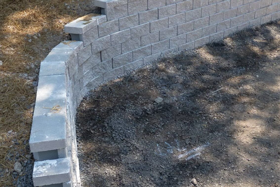 A curved retaining wall made of stacked light gray rectangular concrete blocks built against a sloped dirt bank.