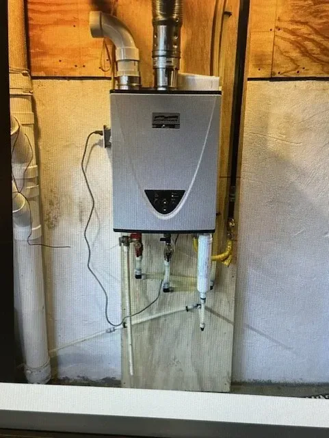 A wall-mounted, silver tankless water heater with vents and pipes, mounted on a wood panel in a basement.