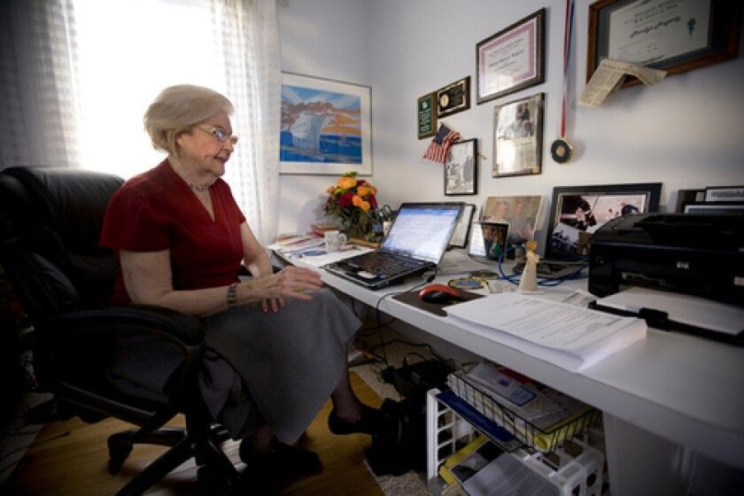 Older woman at a desk, using a laptop. Home office setting with photos, awards, and a printer.
