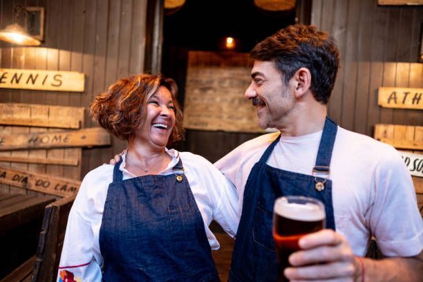 Two people in aprons smiling, embracing, holding a beer in a rustic interior.