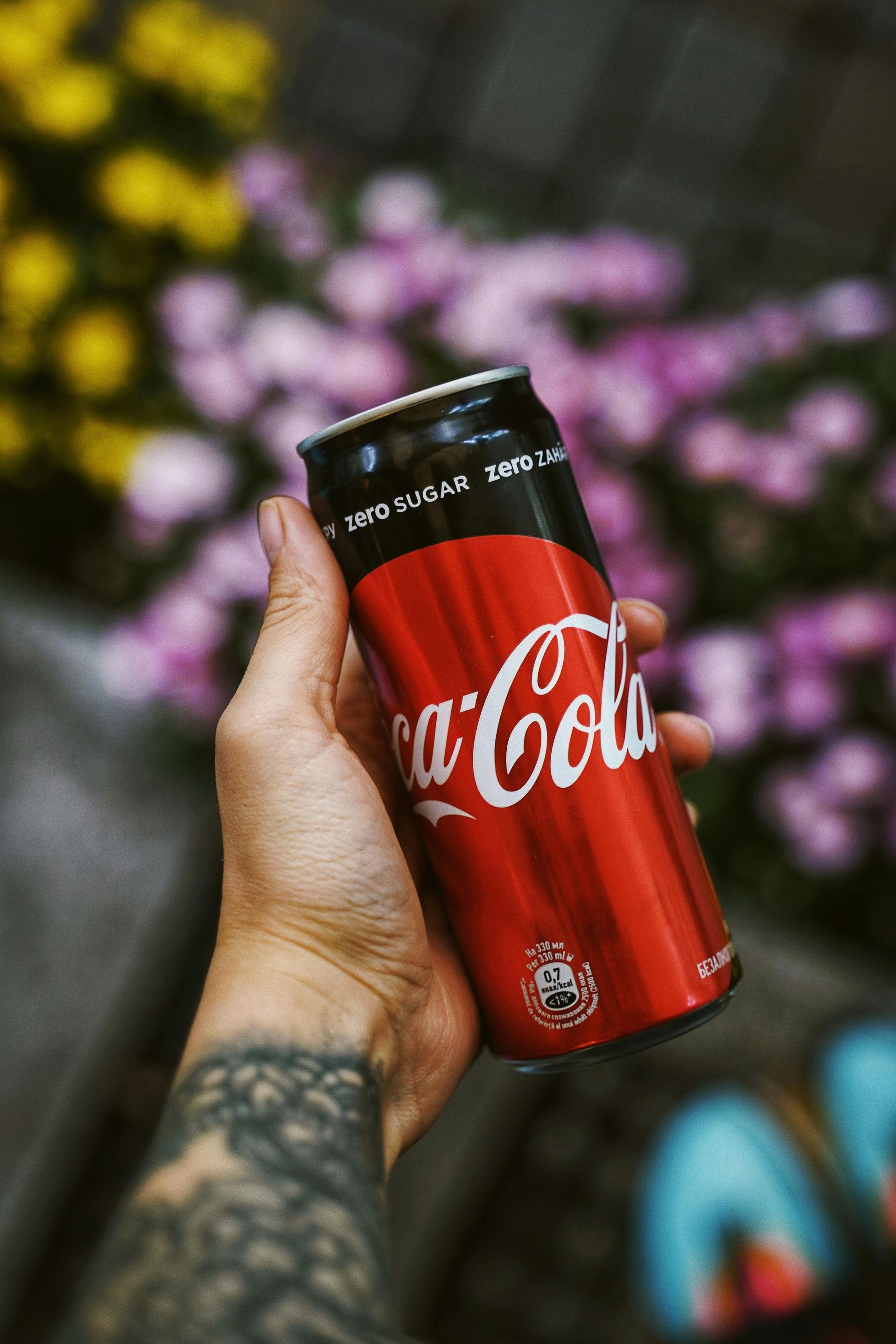 Hand holding a Coca-Cola Zero can in front of flowers.