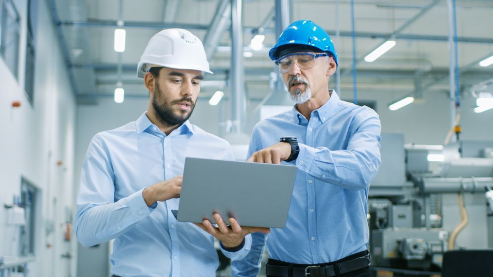 Two men in hard hats are looking at a laptop in a factory.