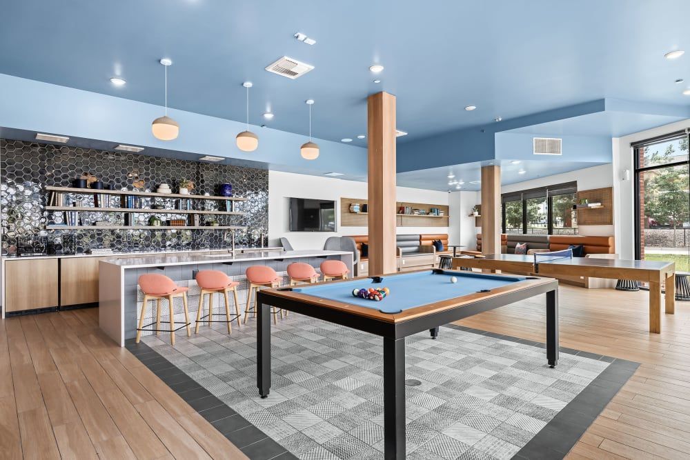 Modern and stylish recreational room with a blue billiard table at the center, bar counter with pink stools, geometric-patterned floor, and wooden accents. The room has a high ceiling with multiple hanging globe lights and large windows providing ample natural light at Marq Iliff Station in Aurora, CO.
