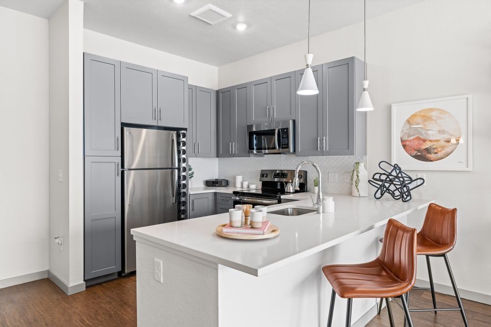 Modern kitchen interior with gray cabinets, stainless steel appliances, and a white countertop island with brown leather stools. Pendant lights hang above, and a decorative abstract sculpture sits on the counter. Artwork hangs on the right wall at Marq Iliff Station in Aurora, CO.